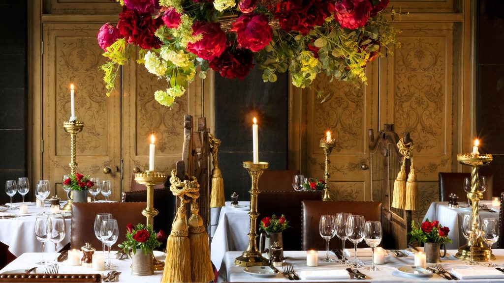 Elegant restaurant dining room with tables set for service, gold candelabras, white tablecloths, wine glasses, and a large hanging floral arrangement with red and green flowers.