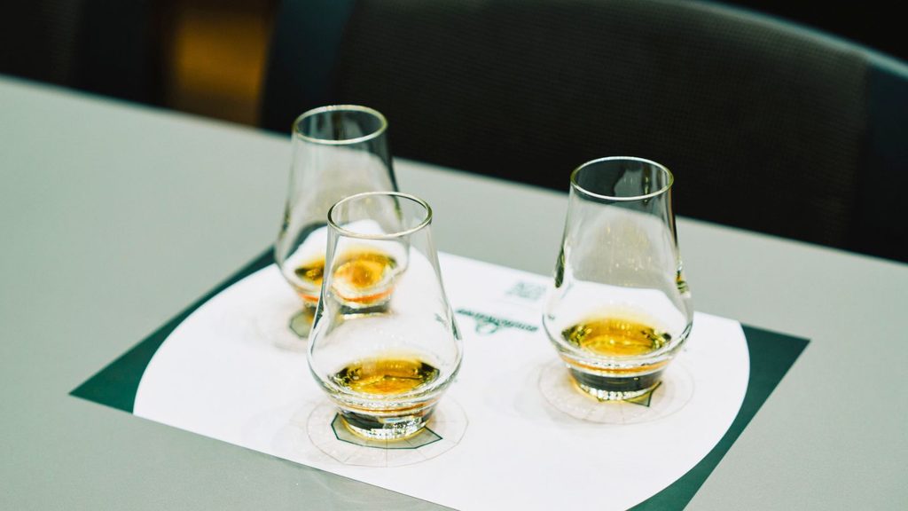 Three glasses with a small amount of amber liquid—likely bourbon and rye—are placed on a white paper mat on a gray tabletop, possibly for a tasting event.