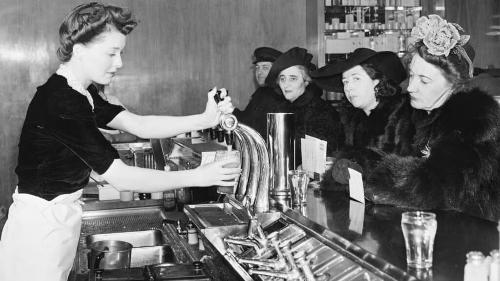 A waitress pours a drink at a counter for three women in hats and fur coats seated at a diner, evoking classic drugstore soda fountain traditions, with menus and glasses visible.