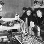 A waitress pours a drink at a counter for three women in hats and fur coats seated at a diner, evoking classic drugstore soda fountain traditions, with menus and glasses visible.