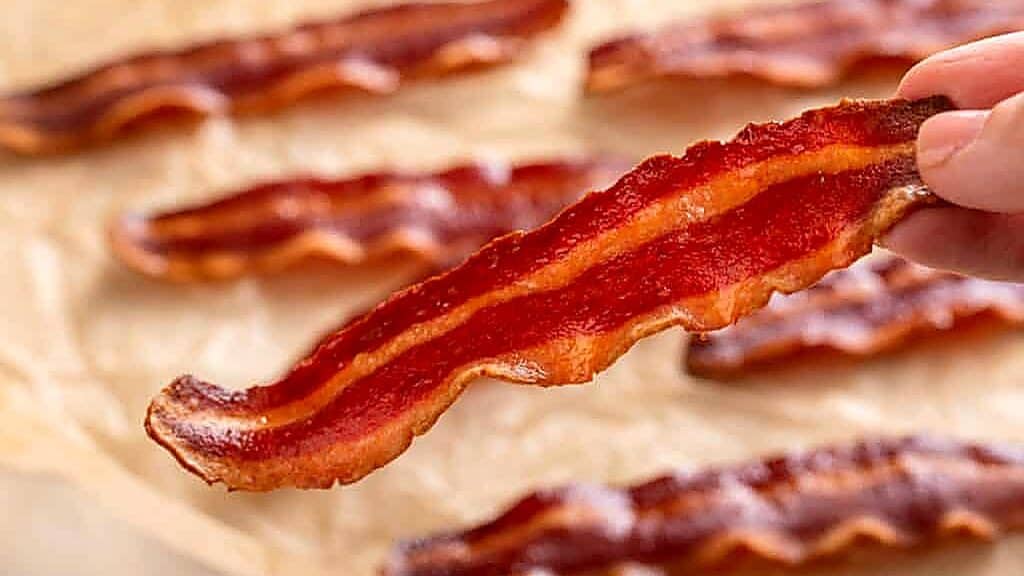 A hand holds a crispy strip of cooked bacon above a surface lined with several other bacon strips on parchment paper, highlighting the delicious results you can achieve&mdash;even when experimenting with ingredient substitutions.