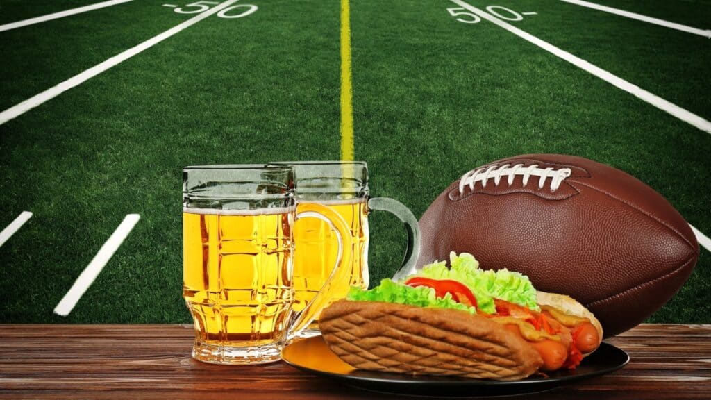 A football, two mugs of beer, and a hot dog on a plate are on a table in front of a football field background, evoking the classic game-day experience found at NFL stadiums with priciest concessions.