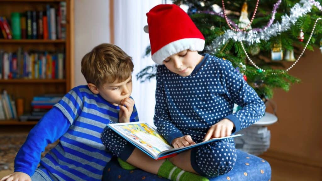 Two children sit by a decorated Christmas tree, one in a Santa hat and pajamas reads a book aloud while the other listens intently—capturing the kind of holiday magic everyone remembers from paging through the Sears Wish Book.