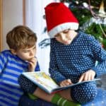 Two children sit by a decorated Christmas tree, one in a Santa hat and pajamas reads a book aloud while the other listens intently&mdash;capturing the kind of holiday magic everyone remembers from paging through the Sears Wish Book.