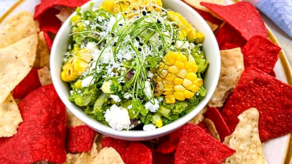 A bowl of guacamole topped with corn, cheese, and sprouts is surrounded by red and yellow tortilla chips on a plate.
