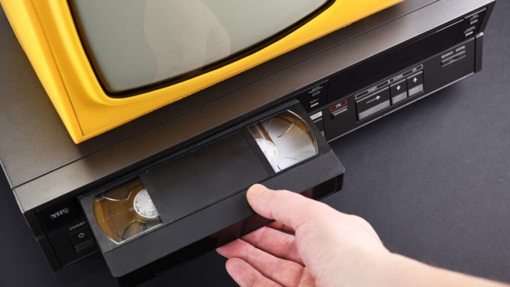 A hand inserts a VHS tape of taped shows on VHS into a VCR, next to a yellow-framed television, all placed on a black surface.