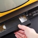 A hand inserts a VHS tape of taped shows on VHS into a VCR, next to a yellow-framed television, all placed on a black surface.