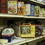 Three shelves display an assortment of vintage metal lunchboxes, including classic 80s lunchbox designs with various motifs, along with some glass bottles, dishes, and kitchenware on the lower shelf.
