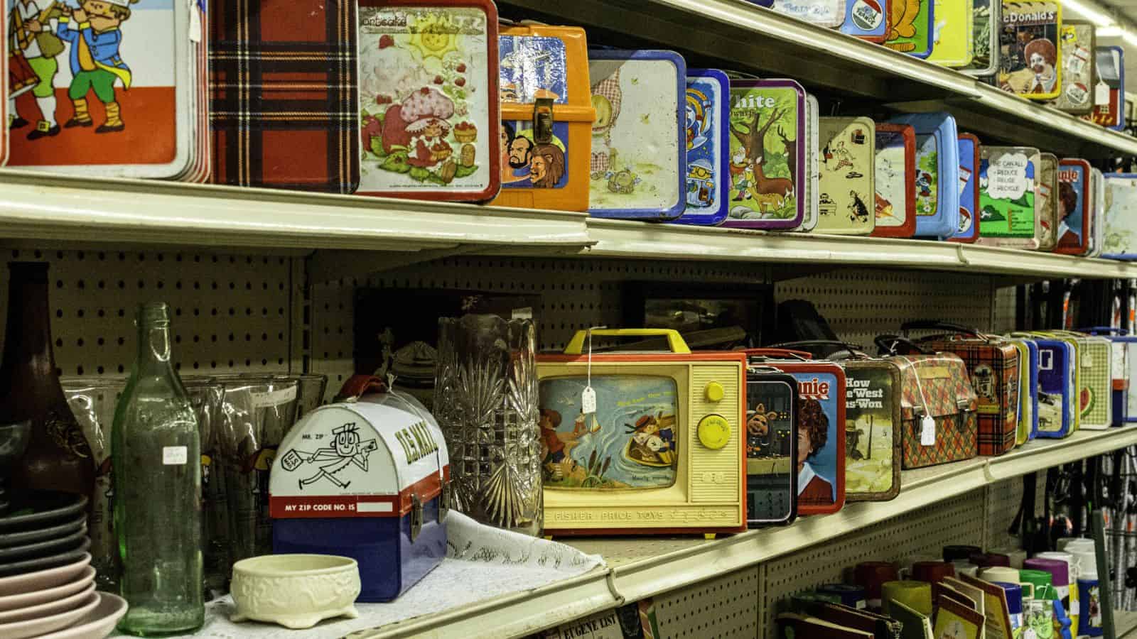 Three shelves display an assortment of vintage metal lunchboxes, including classic 80s lunchbox designs with various motifs, along with some glass bottles, dishes, and kitchenware on the lower shelf.