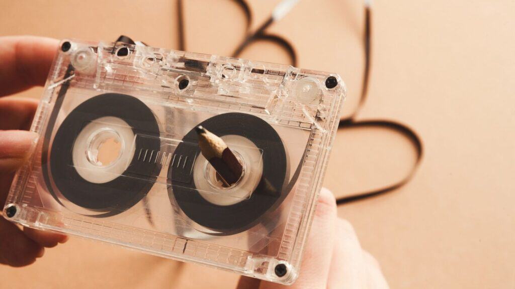 Close-up of hands rewinding a cassette tape with a pencil, evoking classic mixtape making teenager rituals, with some of the tape unraveled in the background.