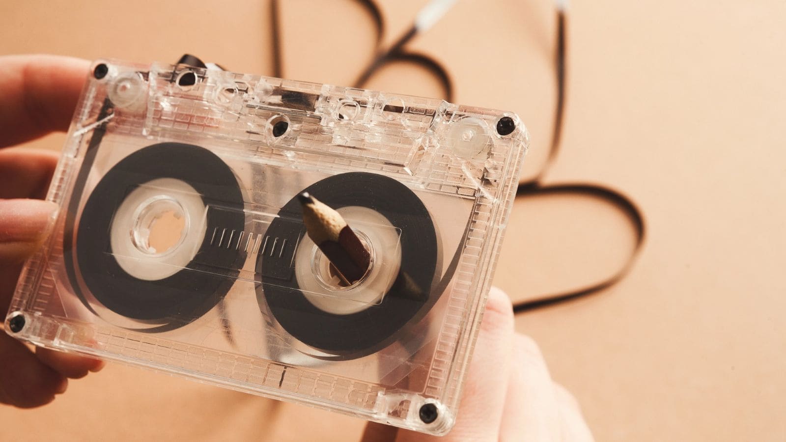 Close-up of hands rewinding a cassette tape with a pencil, evoking classic mixtape making teenager rituals, with some of the tape unraveled in the background.