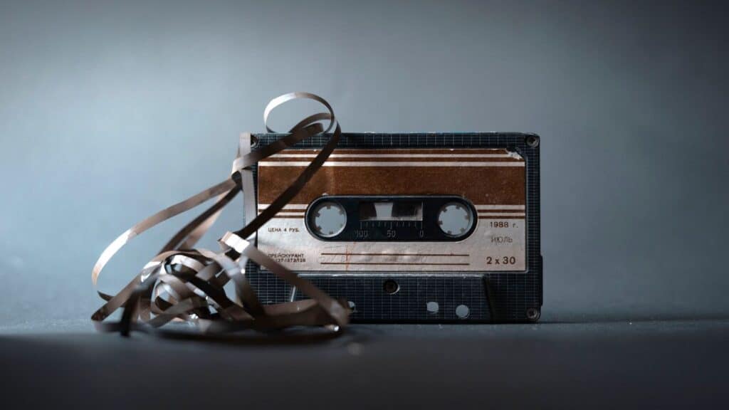 A vintage audio cassette with tangled magnetic tape sits on a dark surface against a gray background, evoking memories of taped shows on VHS and the analog era of home recording.