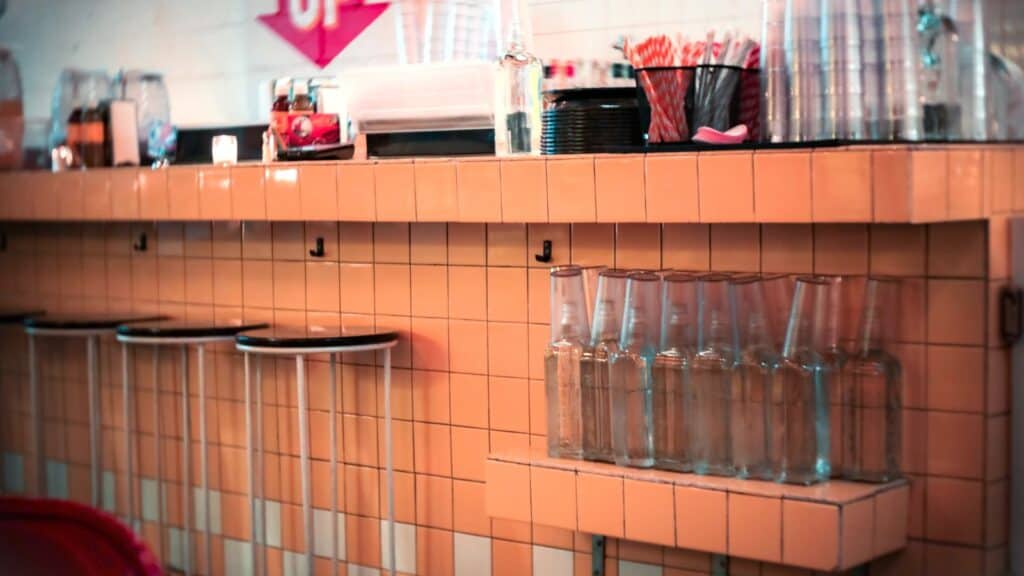 A tiled counter with water bottles, stacked cups, straws, and napkin holders recalls classic drugstore soda fountain traditions; three wall-mounted stools are folded up beneath the counter.