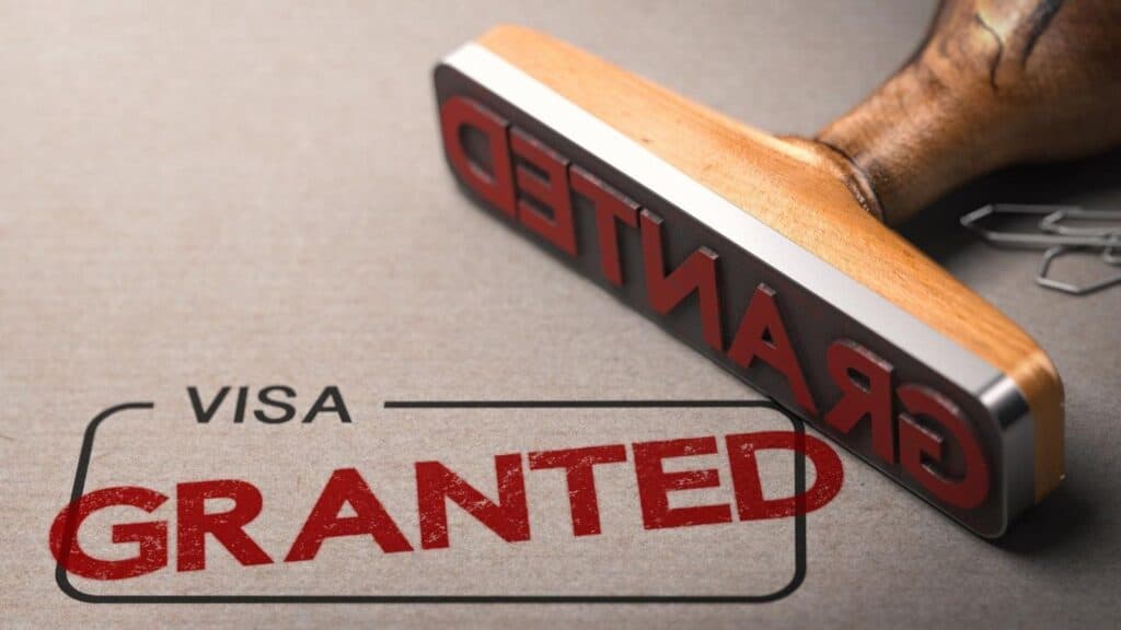 A wooden stamp marks the word &ldquo;GRANTED&rdquo; in red ink below the word &ldquo;VISA&rdquo; on a gray surface, emphasizing that US tourists need approval first.