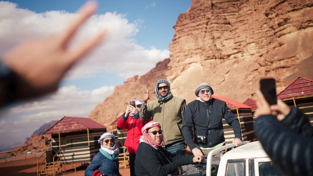 Five people pose for a photo near rocky cliffs and huts in a desert area, while a hand in the foreground displays a peace sign.