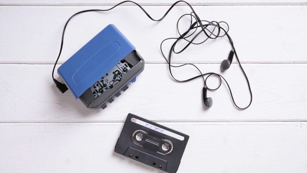 A blue portable cassette player with attached black earbuds and a black audio cassette tape rests on a white wooden surface, evoking memories of when a Walkman owner remembers listening to music on the go.