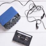 A blue portable cassette player with attached black earbuds and a black audio cassette tape rests on a white wooden surface, evoking memories of when a Walkman owner remembers listening to music on the go.