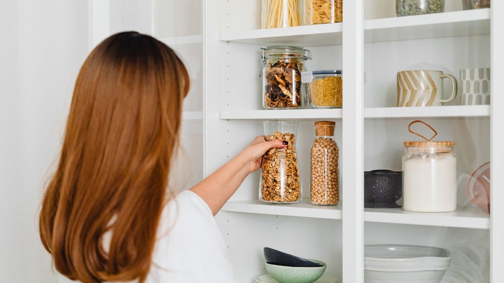A woman with long hair places a glass jar of dried food—one of her essential dorm pantry staples—on a white kitchen shelf filled with jars, mugs, and bowls.