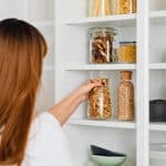 A woman with long hair places a glass jar of dried food—one of her essential dorm pantry staples—on a white kitchen shelf filled with jars, mugs, and bowls.