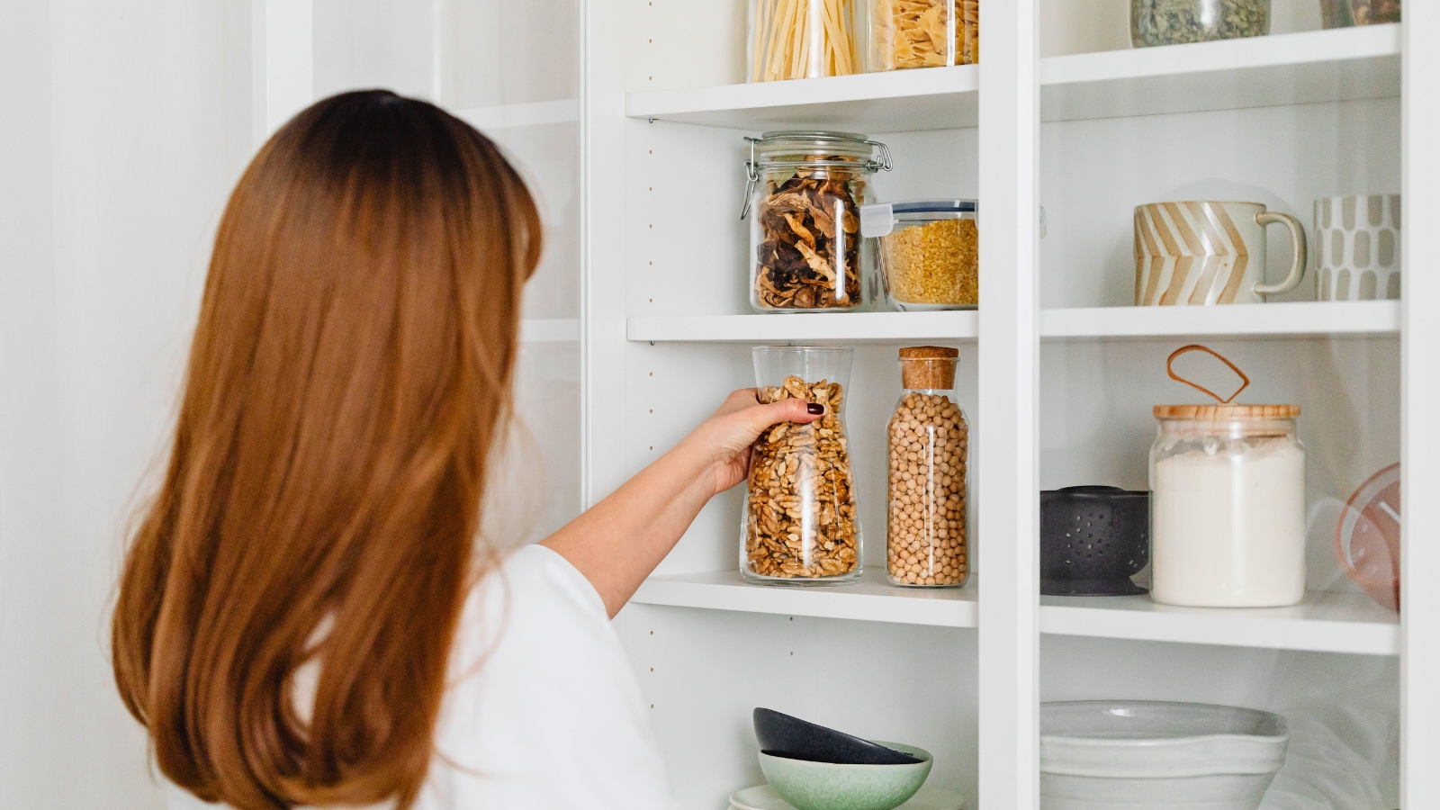 A woman with long hair places a glass jar of dried food—one of her essential dorm pantry staples—on a white kitchen shelf filled with jars, mugs, and bowls.