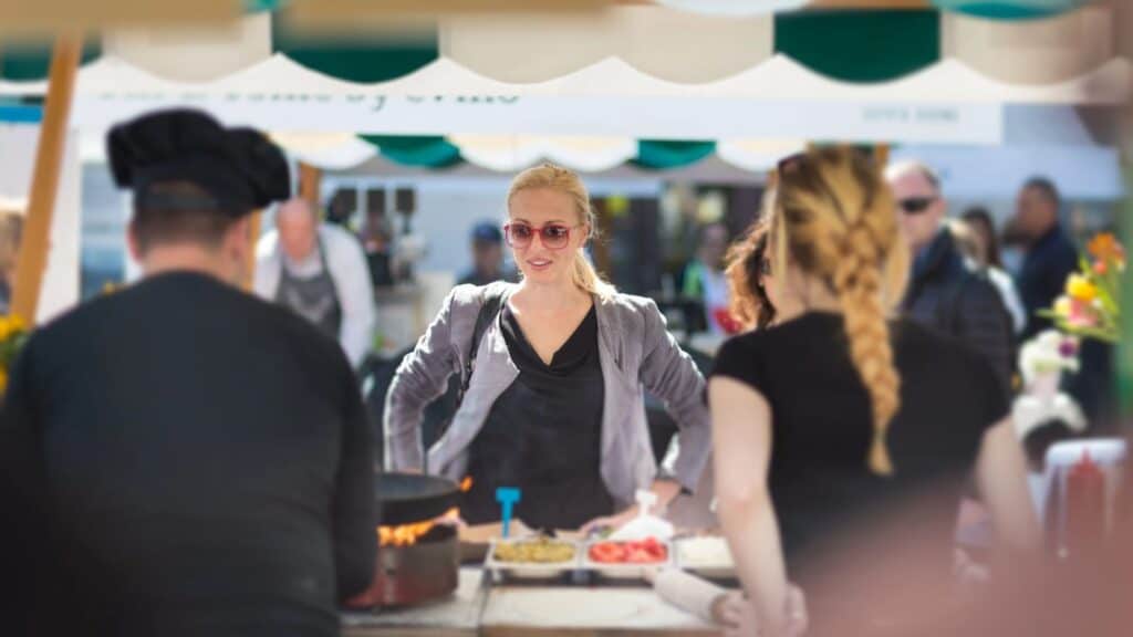 A woman in sunglasses stands behind a food stall counter at an outdoor market, facing two people with their backs to the camera&mdash;a lively scene reminiscent of bustling fall chili cook-offs.