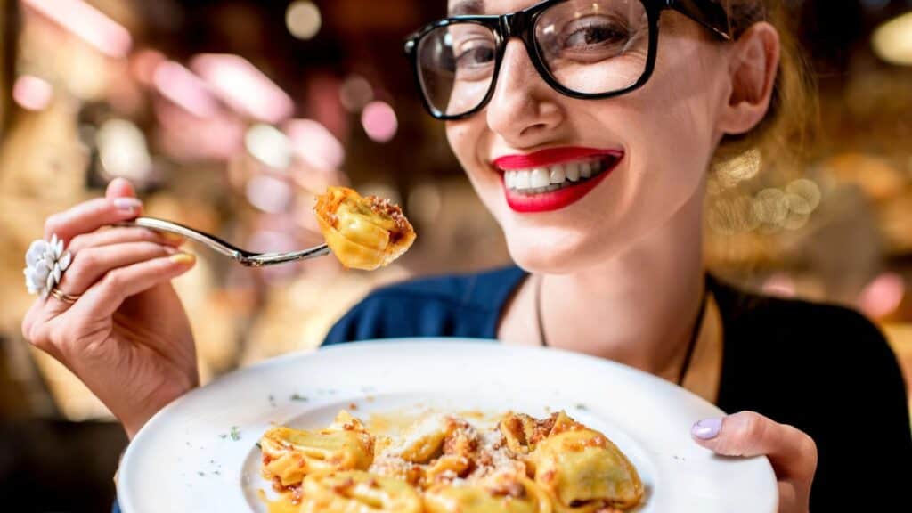 Woman wearing glasses and red lipstick smiling while holding a plate of pasta and lifting a bite with a fork.