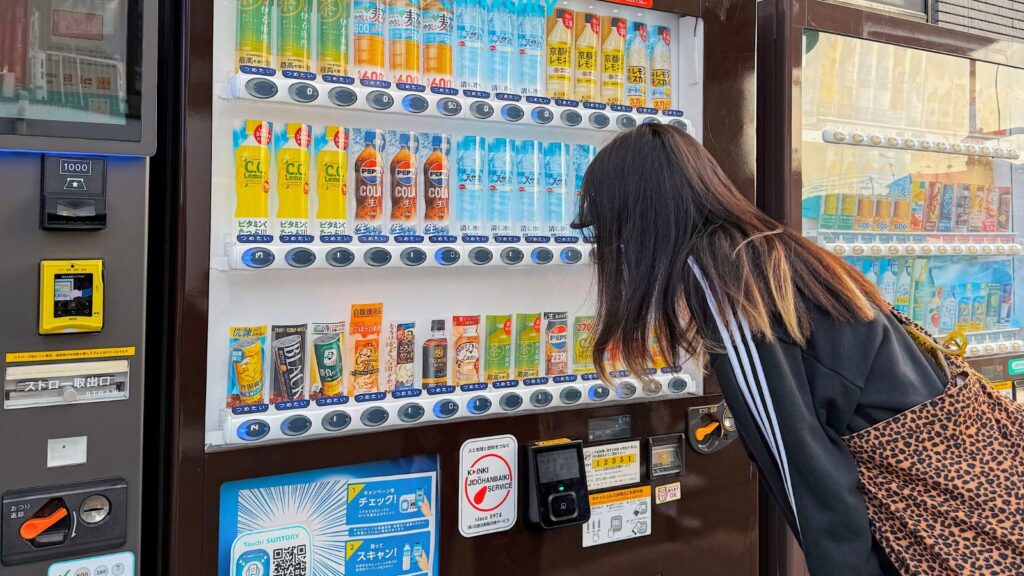 A woman with long hair and a leopard print bag looks closely at a vending machine with various drinks in bottles and cans&mdash;an everyday scene that reflects how the vending machine changed American snacking.