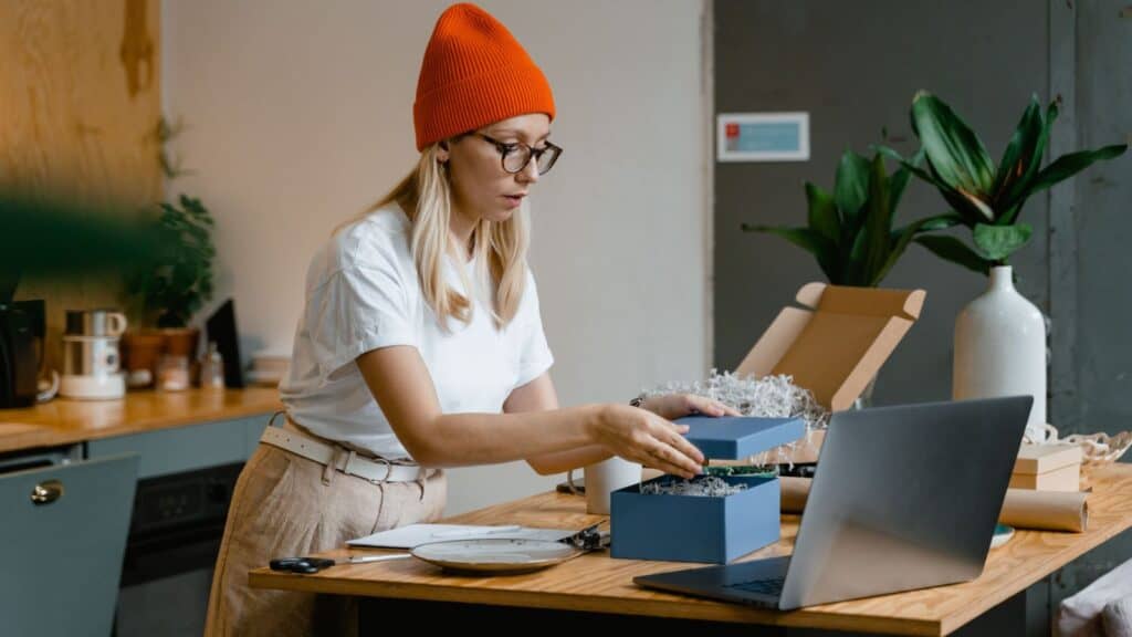 A person in a white shirt and orange hat packs a blue box with shredded paper at a wooden table, with a laptop and packaging materials nearby—busily assembling what could be a thoughtful college care package Co style gift.