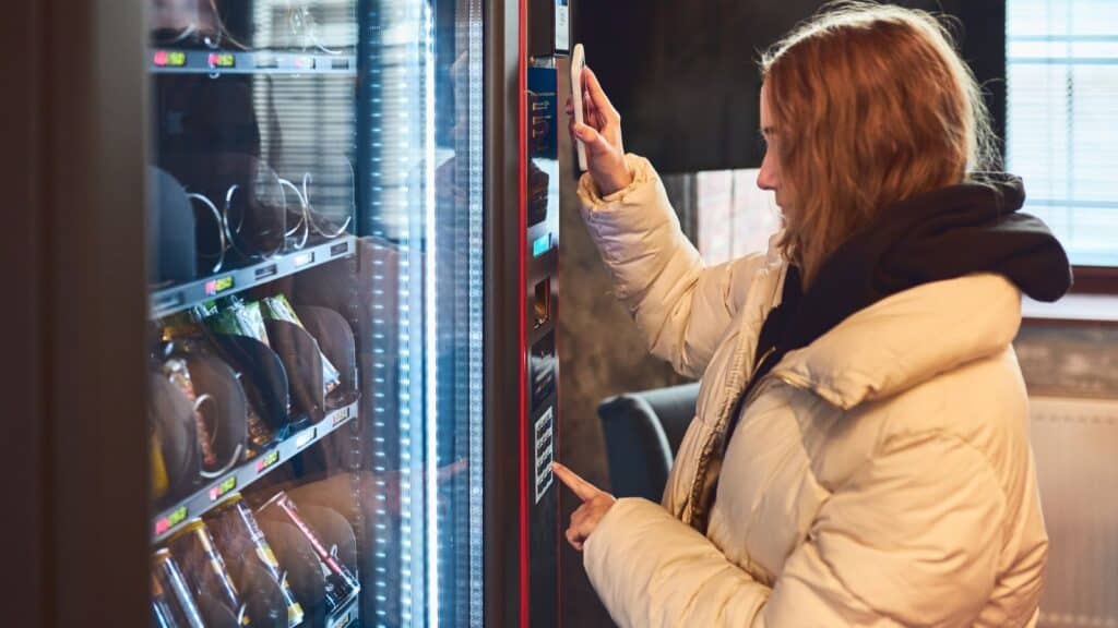 A person in a light-colored puffer jacket uses a vending machine&mdash;one of the innovations that changed American snacking&mdash;pressing buttons and interacting with the payment panel.