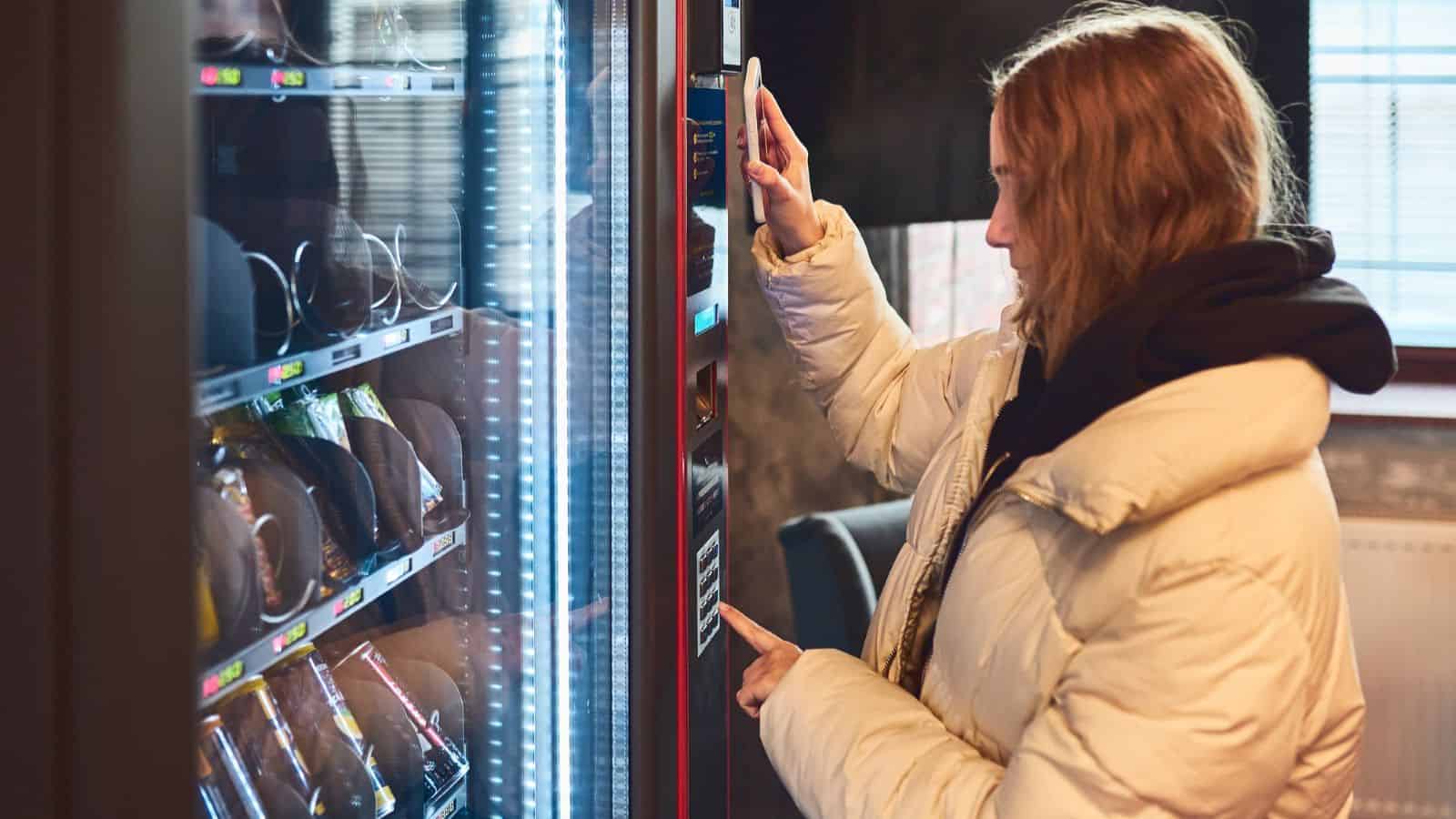 A person in a light-colored puffer jacket uses a vending machine—one of the innovations that changed American snacking—pressing buttons and interacting with the payment panel.