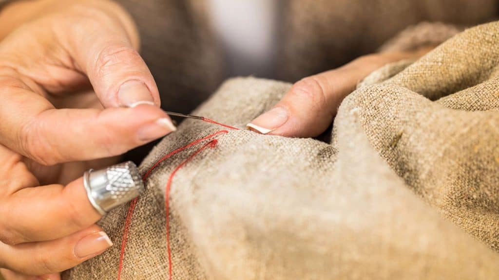 Close-up of hands sewing beige fabric with a needle and red thread, using a metal thimble on one finger.