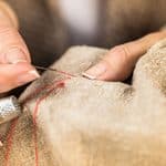 Close-up of hands sewing beige fabric with a needle and red thread, using a metal thimble on one finger.