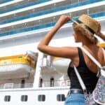 Person with a backpack and hat stands in front of a large white cruise ship with yellow lifeboats.