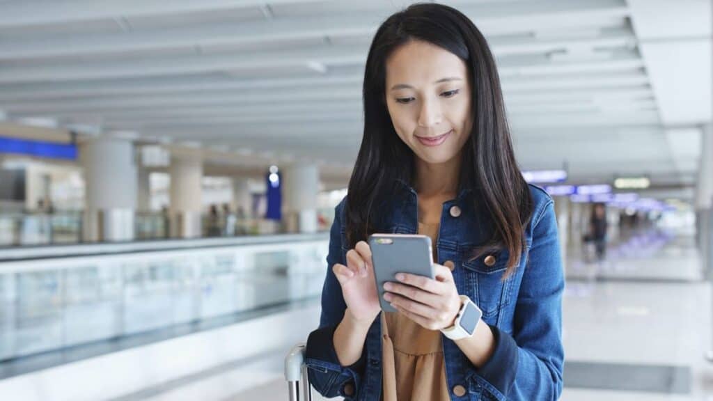 A woman stands in an airport terminal, looking at her smartphone and smiling, with a suitcase next to her&mdash;perhaps she&rsquo;s checking emergency contacts for international travel before her flight.