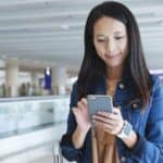 A woman stands in an airport terminal, looking at her smartphone and smiling, with a suitcase next to her—perhaps she’s checking emergency contacts for international travel before her flight.