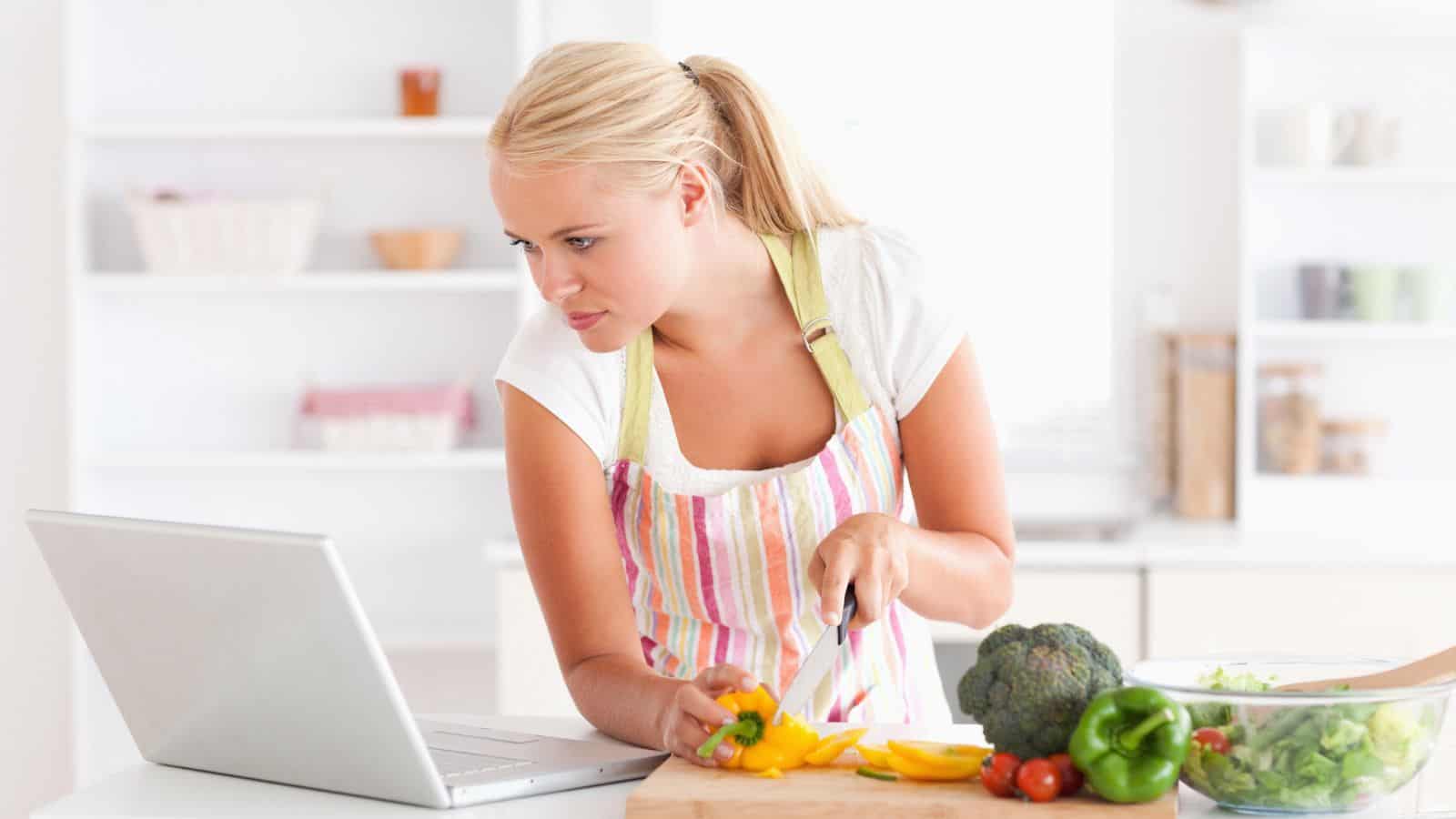 Woman in a striped apron chops vegetables on a cutting board while looking at a laptop in a bright kitchen.