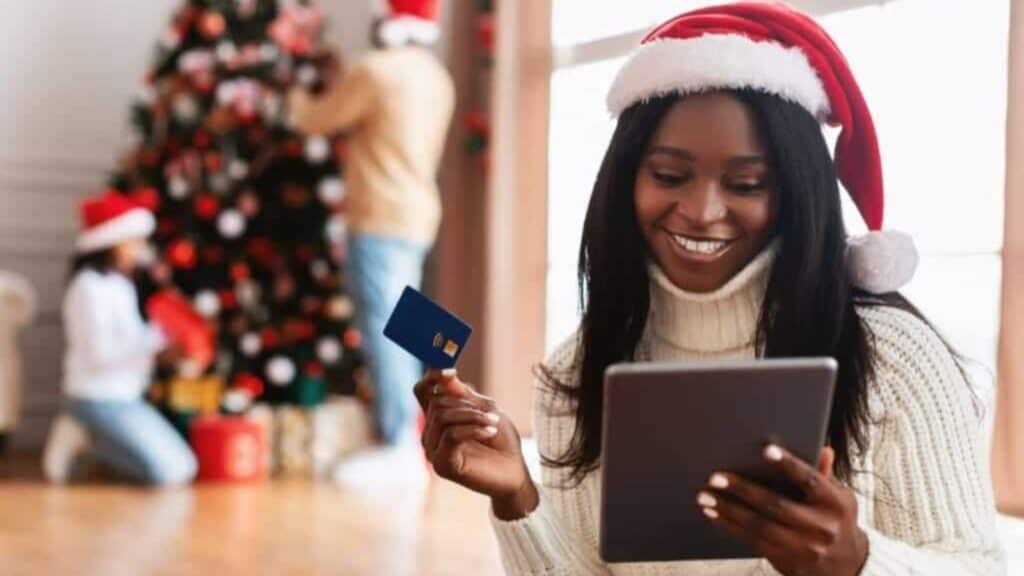 Woman in a Santa hat holds a credit card and tablet, smiling, with a decorated Christmas tree and two people in the background—just the kind of joyful scene everyone remembers from flipping through the Sears Wish Book during the holidays.