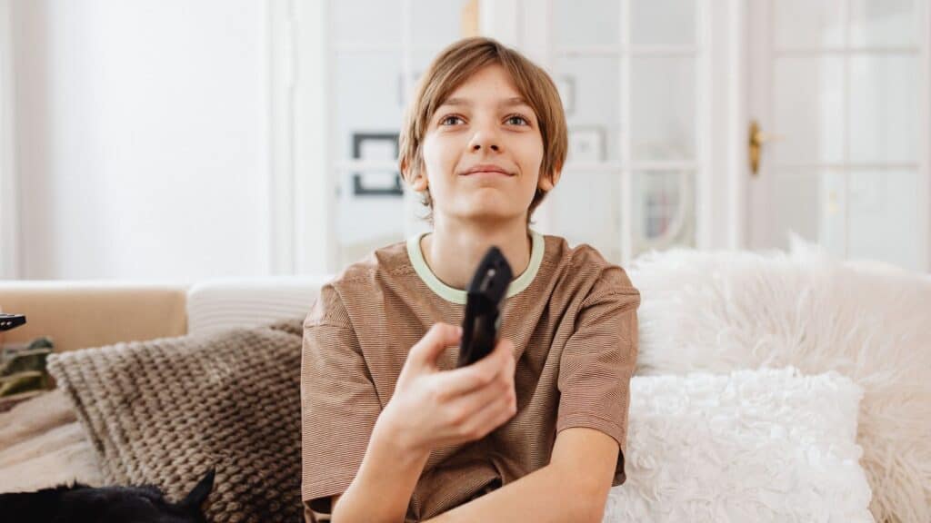 A young person with light brown hair sits on a couch holding a TV remote, looking up and smiling, evoking the joy of Saturday cartoon traditions. There are cushions and a fluffy blanket in the background.