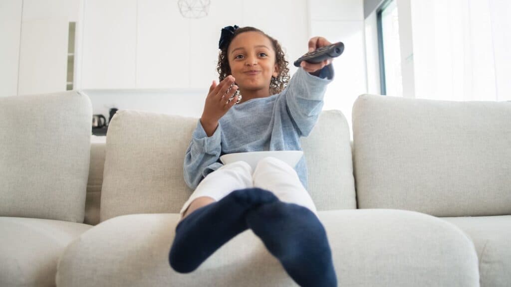 A young girl sits on a white couch, holding a remote control and a bowl of popcorn, enjoying her favorite Saturday cartoon traditions in a bright living room.