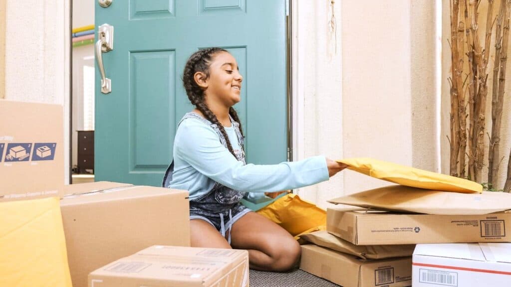 A young girl sits on a doorstep, smiling while sorting through multiple cardboard boxes and yellow padded envelopes—an experience that brings to mind how everyone remembers Sears Wish Book and its promise of surprises delivered right to your door.