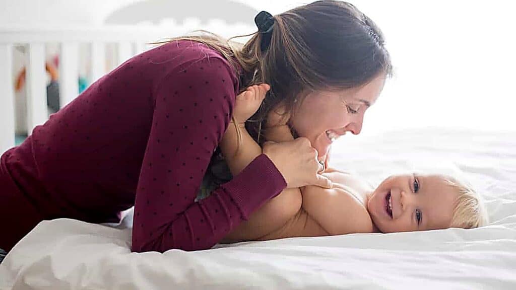 A woman in a maroon shirt smiles while leaning over a laughing baby lying on a white bed.