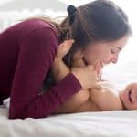 A woman in a maroon shirt smiles while leaning over a laughing baby lying on a white bed.