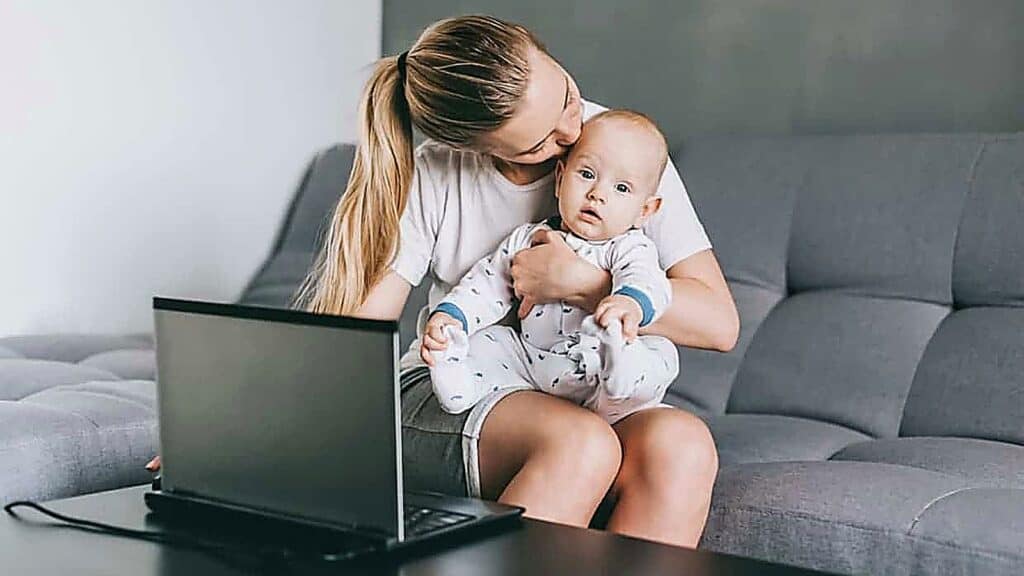 Woman holding a baby on her lap while sitting on a gray sofa next to an open laptop on a table.