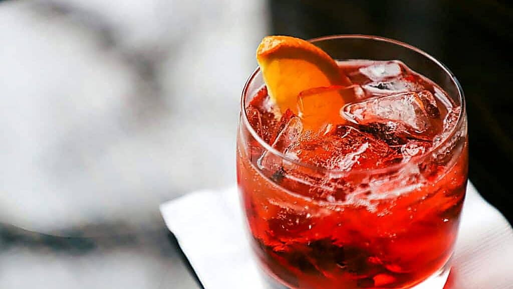A glass of red cocktail with ice and an orange wedge garnish sits on a white napkin, offering a vibrant addition to any selection of vegan drinks.