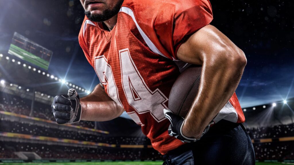 American football player in a red jersey holding a football and running on a stadium field under stadium lights at night.