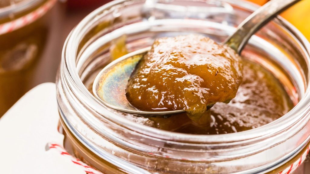 A close-up of a spoon scooping thick brown apple butter from a glass jar, highlighting the rich, velvety texture of the homemade apple butter.