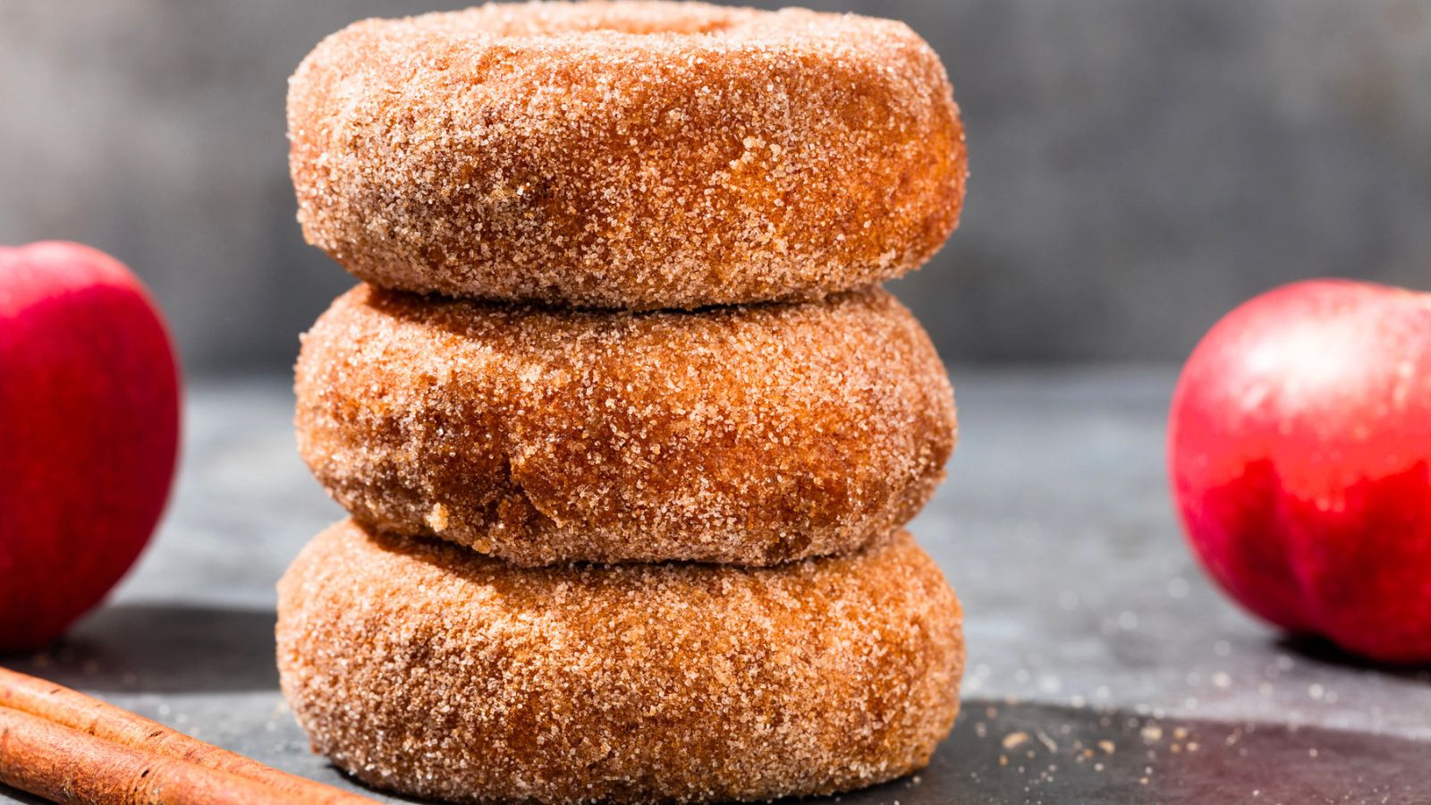Three cinnamon sugar donuts stacked on top of each other, with two red apples and cinnamon sticks placed nearby on a gray surface.