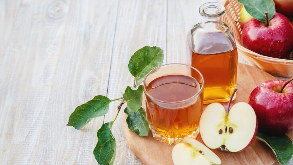 A glass and a bottle of apple juice next to a sliced apple, green leaves, and a basket of red apples on a wooden surface.