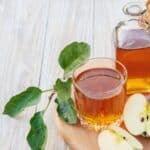 A glass and a bottle of apple juice next to a sliced apple, green leaves, and a basket of red apples on a wooden surface.
