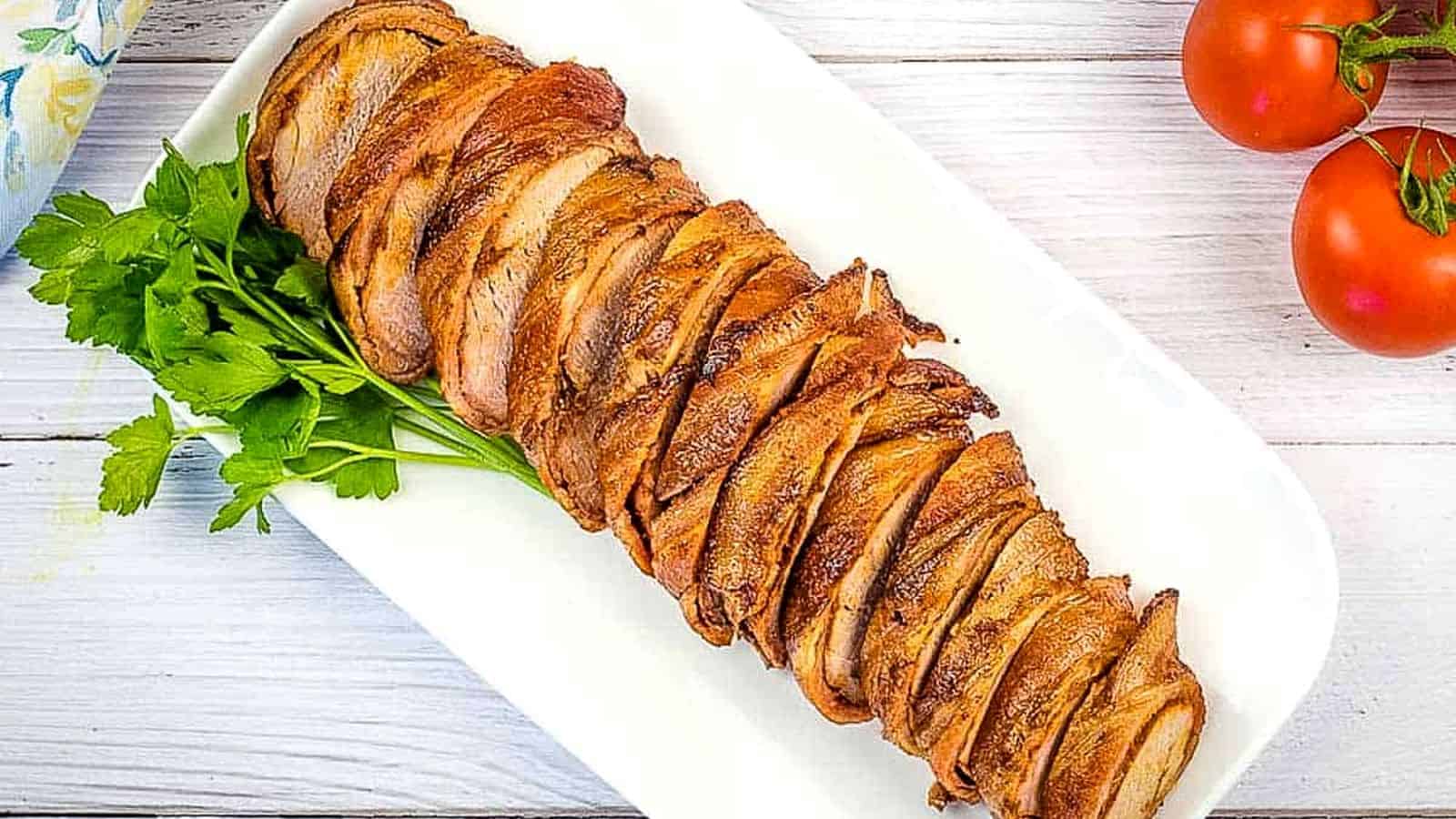 Sliced Smoked Bacon Wrapped Whole Pork Tenderloin on a white platter.